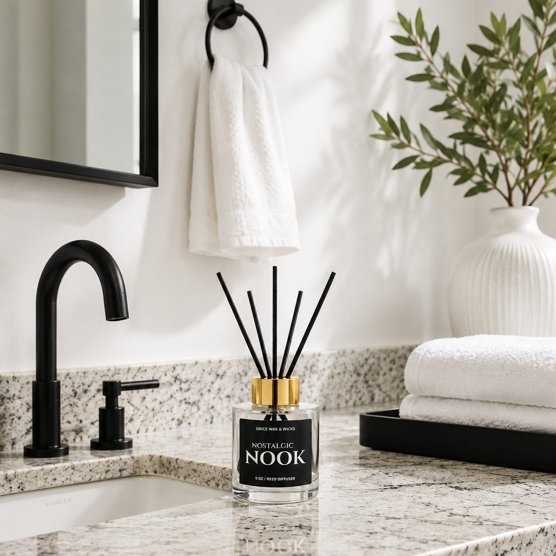 A reed diffuser bottle labeled 'Nook' with black reeds and a gold cap sits on a speckled granite bathroom countertop beside a black faucet, with a white towel, mirror, and plant in the background.