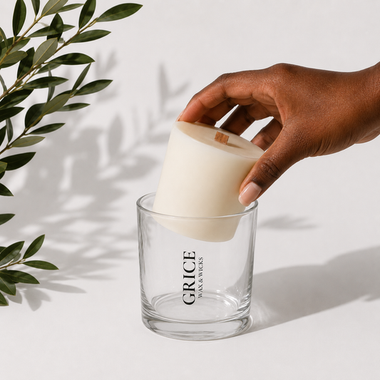 A hand places a wooden wick candle refill into a matte black GRICE Wax & Wicks vessel resting on a neutral linen book titled “The Wax Remembers” against a soft white background with natural light and subtle greenery.