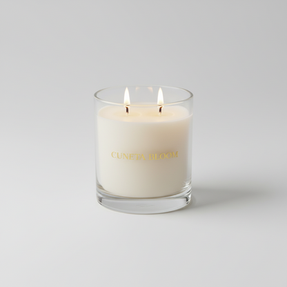 Cuneta Bloom luxury coconut-soy candle in clear glass with gold lettering and double cotton wicks, lit on a neutral cream surface