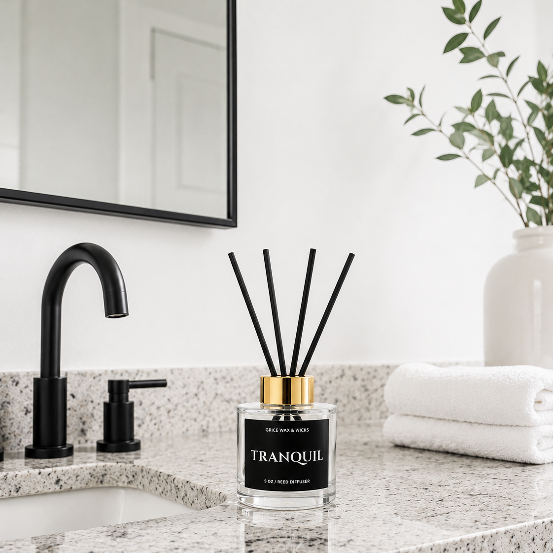 A reed diffuser bottle labeled 'TRANQUIL' sits on a speckled granite bathroom countertop beside a black faucet, with folded white towels and a mirror in the background.