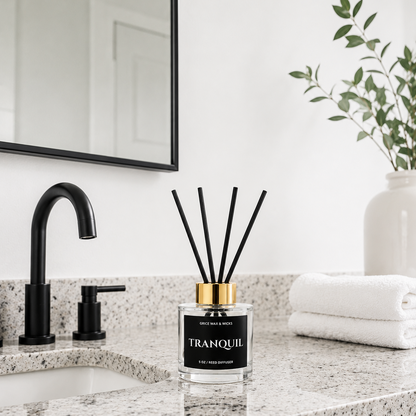 A reed diffuser bottle labeled 'TRANQUIL' sits on a speckled granite bathroom countertop beside a black faucet, with folded white towels and a mirror in the background.